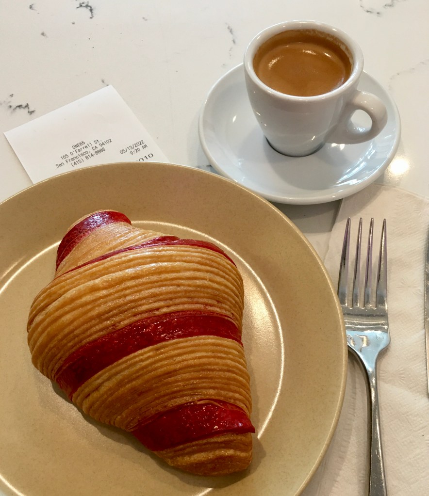 croissant with red raspberry ribbon on brown plate with fork, napkin, espresso in a small espresso cup on a saucer