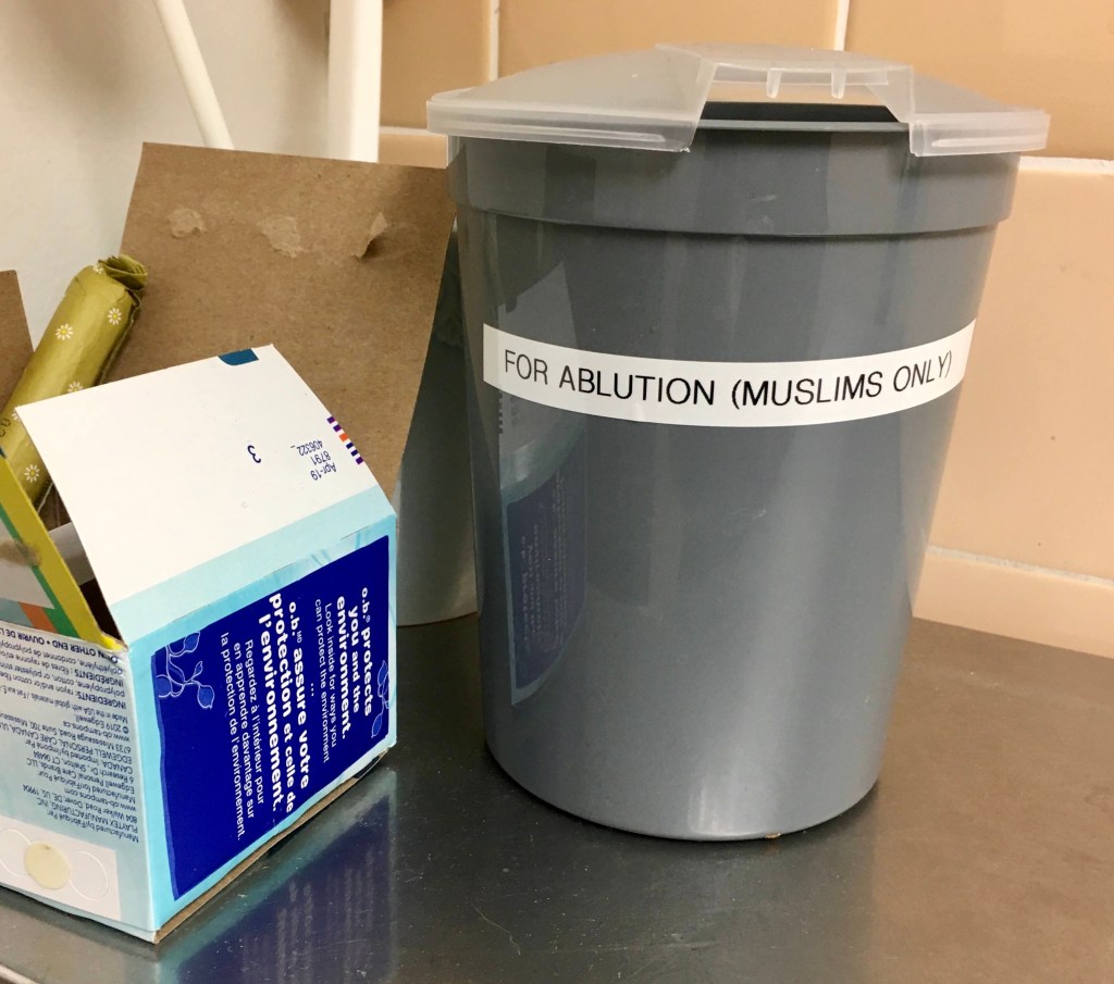 small gray plastic pitcher with a lid with a label that reads “For Ablution (Muslims Only” on public bathroom shelf